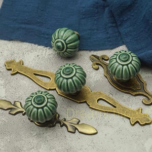 May include: A collection of green ceramic cabinet knobs and pulls with a floral design. The knobs are round and have a ribbed texture. The pulls are brass-colored and feature decorative details, including leaf and scroll designs.