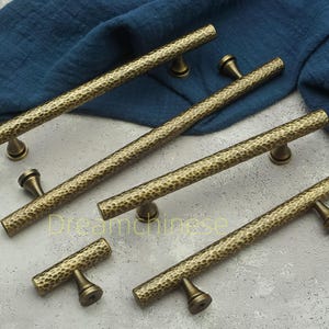 May include: A collection of antique brass cabinet pulls with a hammered texture. The pulls are in various sizes, with a cylindrical bar and rounded mounting posts. The finish is a warm, aged brass color, suggesting a vintage aesthetic.