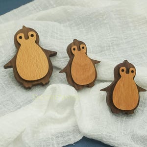 May include: Three wooden penguin figurines with a two-tone design. The outer layer is a dark brown, while the inner body is a lighter wood. The penguins are lying on a white, textured fabric.