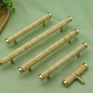 Op de afbeelding: Een verzameling geweven rotan kastgrepen met messing accenten. De grepen zijn cilindrisch met een geweven textuur en messing eindkappen. De hardware is gerangschikt op een lichtgroen oppervlak. Het woord "Dreamchase" is zichtbaar.