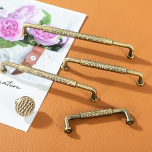 May include: A collection of antique brass cabinet hardware, including three pull handles and one knob. The handles have a hammered texture in the center and are mounted on a brown surface. A magazine with pink roses is in the background.