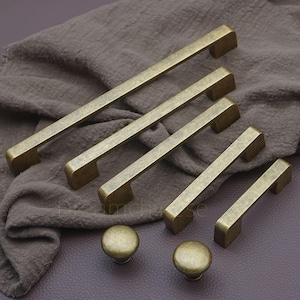 Op de afbeelding: Een verzameling antieke messing kastbeslagen, waaronder verschillende rechthoekige stanggrepen en twee ronde knoppen. De hardware heeft een verouderde afwerking, waardoor het een vintage uitstraling heeft. De grepen variëren in lengte.