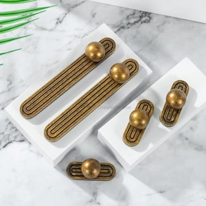 以下が含まれることがあります： アンティーク真鍮製のキャビネット金具のコレクション。長い引き手と小さなノブが含まれています。 金具はボール型のアクセントと溝のあるデザインが特徴です。 大理石の背景に白いプラットフォームに展示されています。