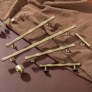 May include: A collection of antique brass cabinet hardware, including several bamboo-style handles and a round knob. The hardware is arranged on a brown fabric and leather surface, showcasing the warm, aged finish of the metal.