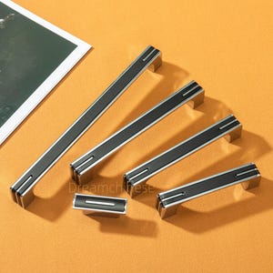 Pode incluir: Um conjunto de puxadores de armário modernos e um botão. O hardware apresenta uma faixa central preta com detalhes prateados. Os puxadores variam em tamanho, medindo o maior cerca de 30 cm. A palavra "Dreamchinese" está impressa na superfície.