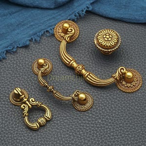 May include: A collection of antique-style brass cabinet hardware, including two handles, a knob, and a pull. The hardware features ornate detailing with floral and scroll designs. The pieces are arranged on a gray surface with a blue cloth in the background.