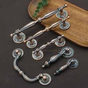 Op de afbeelding: Een assortiment antieke kastknoppen en handgrepen in brons- en kopertinten. De hardware heeft sierlijke details en decoratieve achterplaten. De handgrepen variëren in grootte en vorm, sommige zijn gebogen en andere recht.