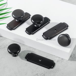 May include: A selection of dark bronze cabinet knobs and pulls. The knobs are circular and the pulls are rectangular with decorative edges. The hardware is displayed on a white surface, highlighting the design and finish.