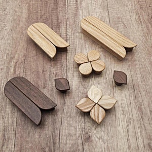 以下が含まれることがあります： さまざまな形と色の木製キャビネットノブとプル。画像には、明るい色の木製プルが2セット、濃い色の木製プルが2セット、葉の形をしたノブが2つ、花の形をしたノブが2つ表示されています。ノブとプルは木製です。