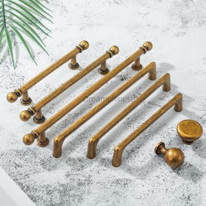 May include: A collection of antique brass cabinet hardware, including several pull handles and two knobs. The pulls vary in length, with a ball-shaped design at each end. The hardware has a vintage, aged appearance.