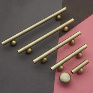 Può includere: Una collezione di ferramenta per mobili color ottone, tra cui maniglie a barra e un pomello. Le maniglie variano in lunghezza, con una barra cilindrica e terminali sferici. Disposti su uno sfondo bicolore grigio e rosa.