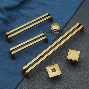 May include: A selection of antique brass cabinet hardware, featuring bar pulls, a round knob, and square knobs. The hardware has a vintage finish and is displayed on a blue and black surface. The text "Dreamchinese" is visible.