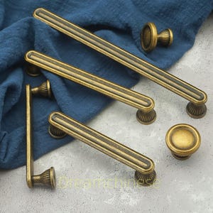 以下が含まれることがあります： アンティーク真鍮のキャビネット金具セット。セットには、長いバープルと丸いノブが含まれています。プルは溝のあるデザインで、装飾的なベースに取り付けられています。ハードウェアは青い布の上に表示されています。