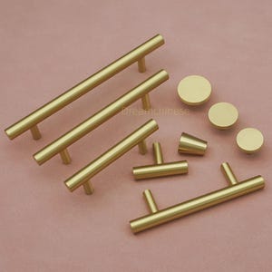 May include: A collection of gold-colored cabinet hardware, including bar pulls, knobs, and a conical handle. The hardware is arranged on a pink surface, showcasing various sizes and styles for furniture upgrades. The hardware is made of metal.