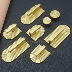 May include: A collection of gold-colored cabinet hardware, including handles and knobs. The handles have a rectangular bar design with a curved base, featuring concentric grooves. The knobs are circular with a spiral pattern. The hardware is arranged on a dark surface.