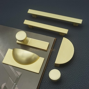 May include: A selection of gold-toned cabinet hardware, including bar pulls, a half-moon pull, and round knobs. The hardware has a ribbed design and is displayed on a neutral surface.