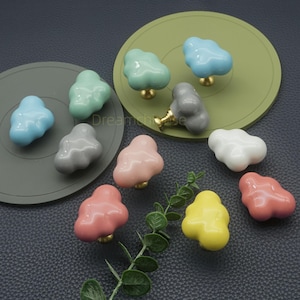Peut inclure: Divers boutons de placard en forme de nuage, de couleurs variées : bleu, vert, rose, gris, blanc et jaune. Chaque bouton est doté d'une base dorée. Les boutons sont disposés sur une surface sombre avec des dessous de plat circulaires verts et gris.