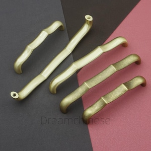 May include: A collection of brass-toned cabinet handles in various sizes. The handles have a bamboo-like design with curved ends and a textured surface. The handles are arranged on a two-tone background of dark gray and maroon.