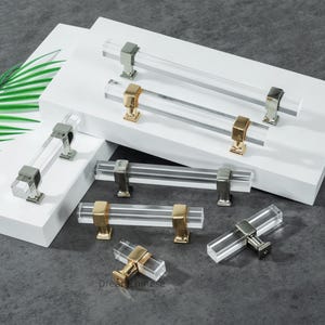 以下が含まれることがあります： 金と銀の金属アクセントが付いた、透明なアクリル製のキャビネットプルコレクション。プルは長方形で、キッチンやバスルームのキャビネットに適したモダンなデザインです。ハードウェアは白い表面に表示されています。