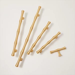 Op de afbeelding: Een verzameling goudkleurige kastgrepen en -knoppen. De hardware heeft een op bamboe ge&iuml;nspireerd ontwerp, met verschillende lengtes en een T-vormige knop. De grepen zijn op een witte achtergrond geplaatst.