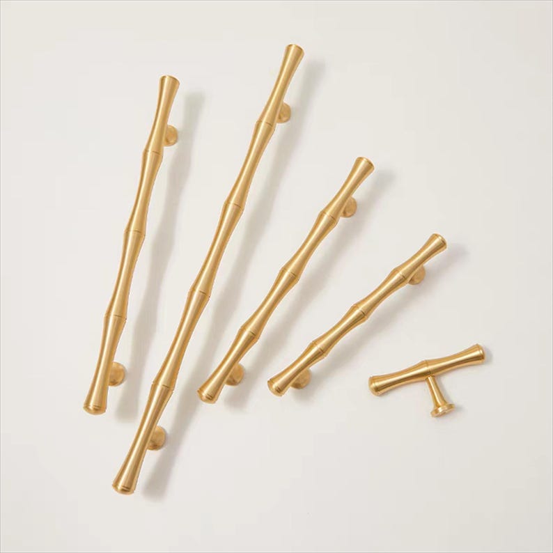 Op de afbeelding: Een verzameling goudkleurige kastgrepen en -knoppen. De hardware heeft een op bamboe ge&iuml;nspireerd ontwerp, met verschillende lengtes en een T-vormige knop. De grepen zijn op een witte achtergrond geplaatst.