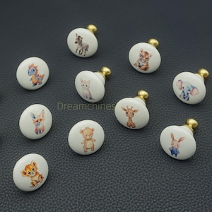 May include: A collection of white ceramic cabinet knobs with gold-colored hardware. Each knob features a different watercolor animal illustration, including a zebra, tiger, dinosaur, cat, fox, bear, lion, giraffe, elephant, and rabbit. These knobs are perfect for adding a whimsical touch to furniture.