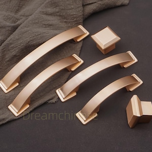 May include: A collection of gold-colored cabinet hardware, including several curved handles and two square knobs. The hardware is arranged on a dark surface with a soft, textured cloth in the background. The word "Dreamchii" is visible.