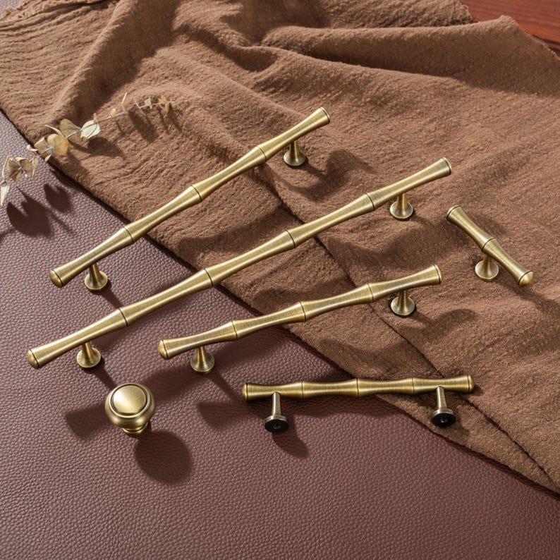 Op de afbeelding: Een verzameling antieke messing kastbeslagen, waaronder verschillende handgrepen in bamboestijl en een ronde knop. De hardware is gerangschikt op een bruine stof en een leren oppervlak, waarbij de warme, verouderde afwerking van het metaal wordt getoond.