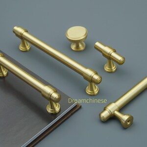May include: A selection of gold-coloured cabinet hardware, including bar pulls, knobs, and t-shaped pulls. The hardware is arranged on a grey surface, with a book visible in the background. The hardware has a matte finish.