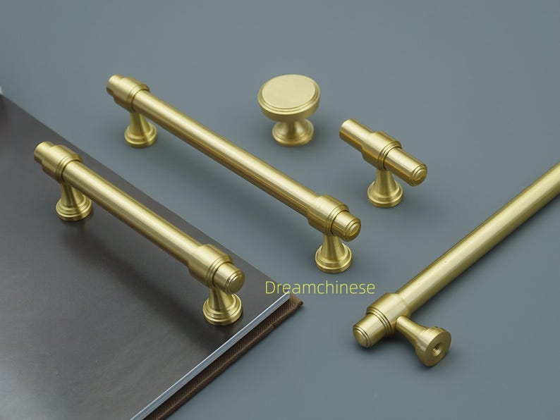 May include: A selection of gold-coloured cabinet hardware, including bar pulls, knobs, and t-shaped pulls. The hardware is arranged on a grey surface, with a book visible in the background. The hardware has a matte finish.