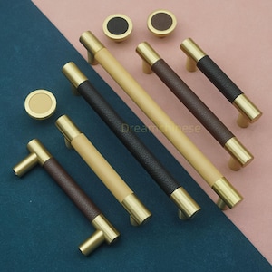 May include: A collection of cabinet hardware, including brass-toned handles and knobs. The handles feature leather-wrapped center sections in brown, black, and tan. The knobs have matching leather inserts. The hardware is arranged on a blue and pink surface.