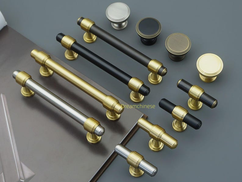 May include: A selection of cabinet handles and knobs in various metallic finishes. The handles are available in black, gold, and silver, with gold detailing. The knobs are round and come in different metallic colours. The hardware is displayed on a grey surface.