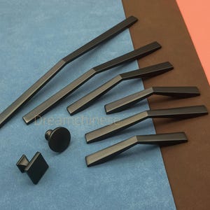 May include: A collection of matte black cabinet hardware, including various sizes of handles and knobs. The selection features long, straight pulls, angled pulls, a round knob, and a square knob. The hardware is displayed on a blue and brown surface.