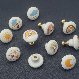 Pode incluir: Uma coleção de puxadores de armário de cerâmica branca com vários desenhos coloridos. Os puxadores apresentam ilustrações de arco-íris, nuvens, estrelas e um sol. Cada puxador tem uma base dourada.