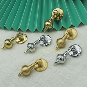 May include: A collection of decorative cabinet pulls in gold and silver finishes. Each pull features a bulbous center, a decorative finial, and a circular backplate. The pulls are arranged on a white surface with a green paper fan in the background.