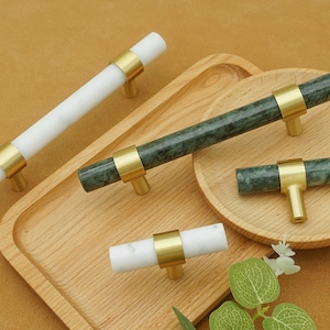 Puede incluir: Herrajes para gabinetes con tiradores de mármol blanco y verde con detalles de latón. Los tiradores están disponibles en varios tamaños y formas, incluidos estilos de barra y tirador en T. El hardware se muestra en una bandeja de madera.