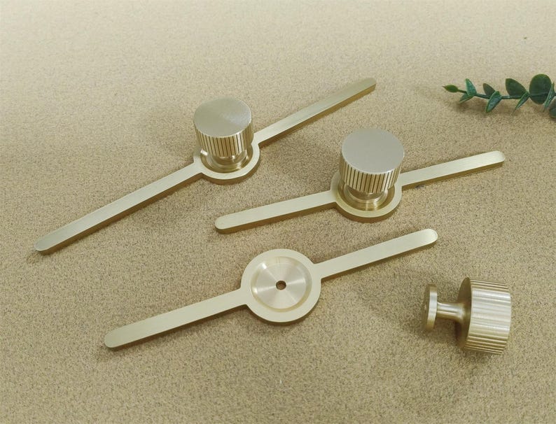 Pu&ograve; includere: Quattro pomelli e maniglie per mobili color ottone. I pomelli hanno una forma cilindrica con dettagli a coste e le maniglie hanno un design lineare. L'hardware &egrave; disposto su una superficie dai toni neutri.