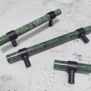 May include: Three cabinet pulls with a green marble effect and black fittings. The cylindrical pulls have a T-shaped design, suitable for drawers or cabinet doors. The marble has a mottled pattern.