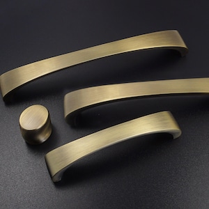 Può includere: Un set di ferramenta per mobili in ottone antico, tra cui tre maniglie a barra curva e un pomello rotondo. La ferramenta ha una finitura spazzolata ed è disposta su una superficie scura. Ideale per armadi.