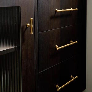 Op de afbeelding: Donkere houten kast met drie lades en een glazen deur. De kast heeft goudkleurige bamboe-stijl handgrepen en een bijpassende deurknop. Het hout heeft een rijke, donkere afwerking.
