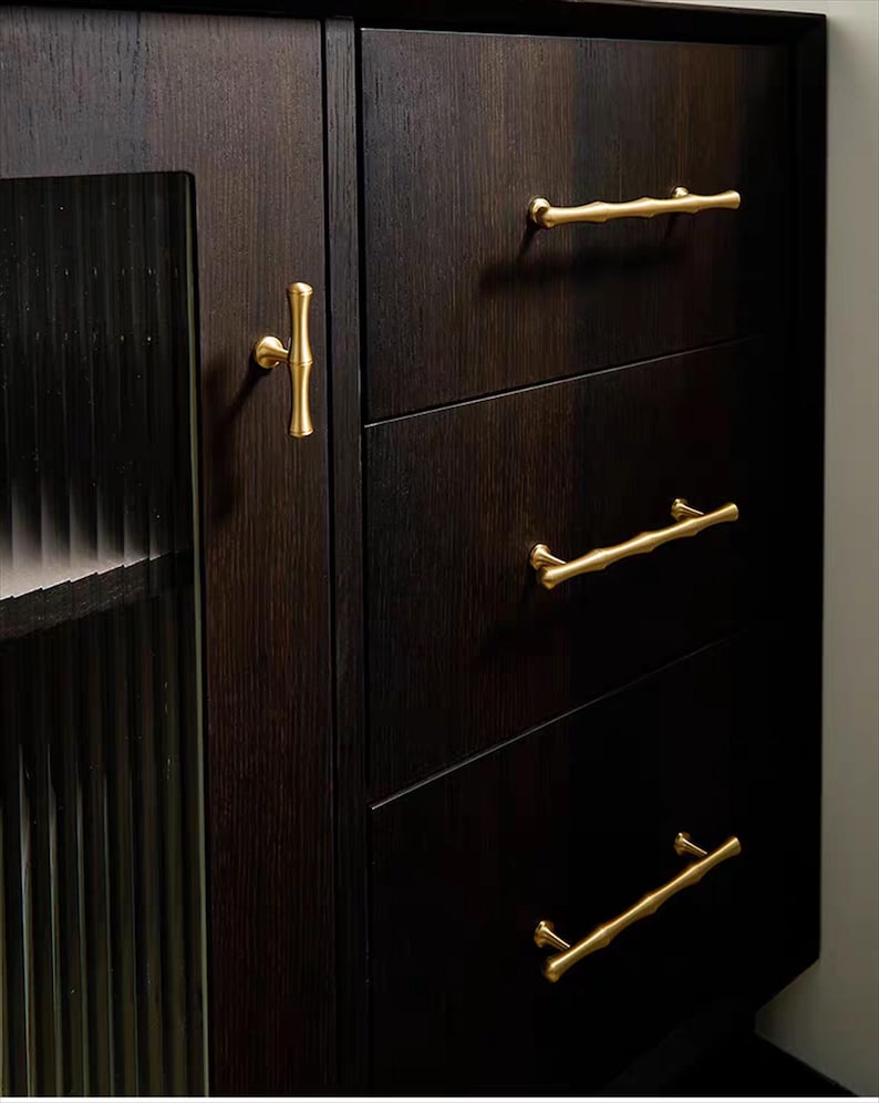Op de afbeelding: Donkere houten kast met drie lades en een glazen deur. De kast heeft goudkleurige bamboe-stijl handgrepen en een bijpassende deurknop. Het hout heeft een rijke, donkere afwerking.