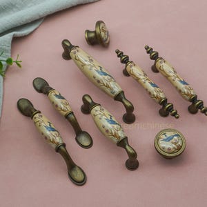 Puede incluir: Un conjunto de tiradores y pomos de armario de estilo antiguo. Los tiradores presentan un centro de cerámica crema con un diseño de pájaro azul y floral, sobre una base de metal bronceado. Los pomos tienen un diseño similar.