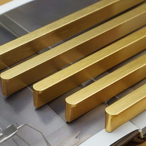 Op de afbeelding: Een set van vijf messing kastgrepen in verschillende lengtes. De rechthoekige grepen hebben afgeronde uiteinden en een geborstelde afwerking. Geschikt voor meubelrenovatie.