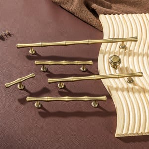 Op de afbeelding: Een verzameling antieke messing kastbeslagen, waaronder handgrepen in verschillende maten en een ronde knop, allemaal in bamboestijl. Gepresenteerd op een bruin oppervlak met een houten accent, waarbij het ontwerp en de afwerking worden benadrukt.