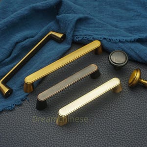 Könnte beinhalten: Eine Auswahl an Schrankbeschlägen in verschiedenen Metallausführungen. Die Auswahl umfasst Stangengriffe, einen Knopf und einen Muschelgriff. Farben sind Gold, Bronze und Schwarz. Die Hardware wird auf einer dunklen Oberfläche mit blauem Stoffhintergrund präsentiert.