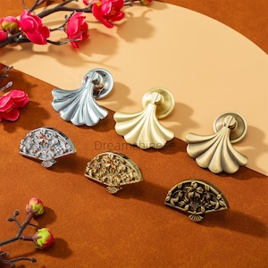 May include: A selection of cabinet hardware in silver, gold, and bronze finishes. The image displays knobs and pulls with fan and draped designs. The hardware is set against a brown background, with red flowers and buds adding a decorative touch.