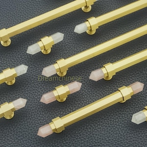 Pode incluir: Close-up de várias puxadores de armário dourados com detalhes em cristal. Os puxadores têm um design de barra retangular com um cristal facetado no centro. Alguns cristais são transparentes, outros são rosa claro. O hardware está em um fundo escuro.