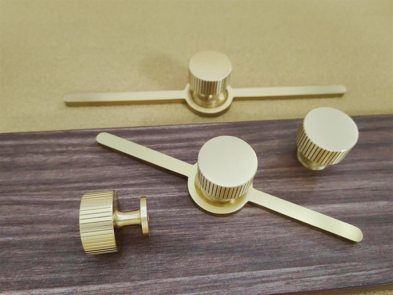 Pu&ograve; includere: Pomelli e maniglie dorate per mobili. I pomelli hanno una forma cilindrica con scanalature verticali e le maniglie hanno un pomello circolare con una barra piatta. L'hardware &egrave; esposto su una superficie di legno scuro.