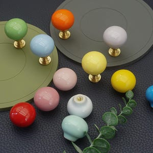 Könnte beinhalten: Eine Sammlung farbenfroher Keramik-Schrankknöpfe in verschiedenen Farbtönen, darunter Grün, Orange, Blau, Rosa und Gelb. Jeder Knopf hat eine runde Form und ist auf einer Messingbasis montiert. Die Knöpfe sind auf einer dunklen Oberfläche angeordnet.