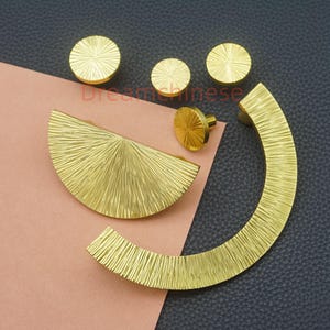 May include: A collection of gold-colored cabinet hardware, including knobs and handles. The hardware features a textured, radial design. The set includes circular knobs, a half-circle handle, and a curved handle. The hardware is displayed on a pink and black surface.